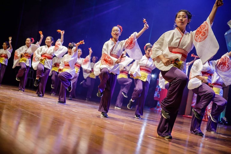Bailarinas con trajes blancos, rojos y morados, bailando animadamente en un escenario iluminado, sosteniendo palillos y elementos de percusión.