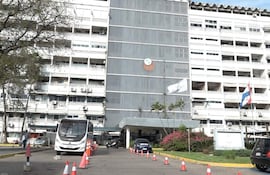 Edificio administrativo del IPS con autobús y automóvil estacionados, banderas y conos de tráfico visibles.