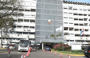 Edificio administrativo del IPS con autobús y automóvil estacionados, banderas y conos de tráfico visibles.