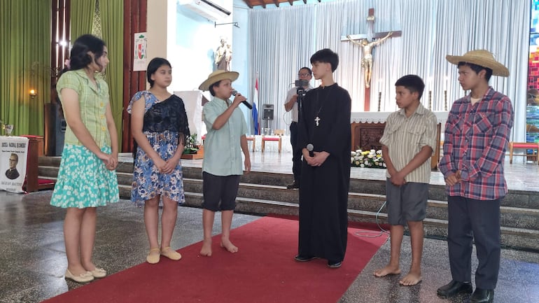 Seis niños participan en una representación teatral, uno sostiene un micrófono vestido de paja, mientras los demás observan atentos en un ambiente escolar.