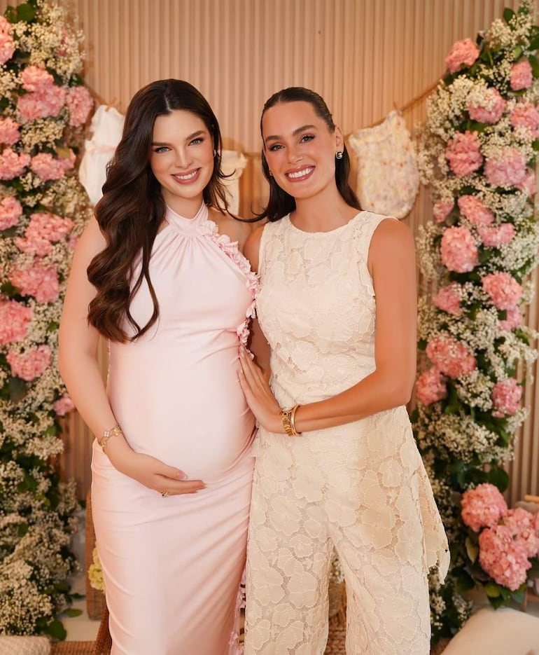 Nadia Ferreira en vestido rosa, tocándose la barriga, sonríe junto a Stephi Stegman en conjunto blanco, en decoración de flores rosadas y blancas.