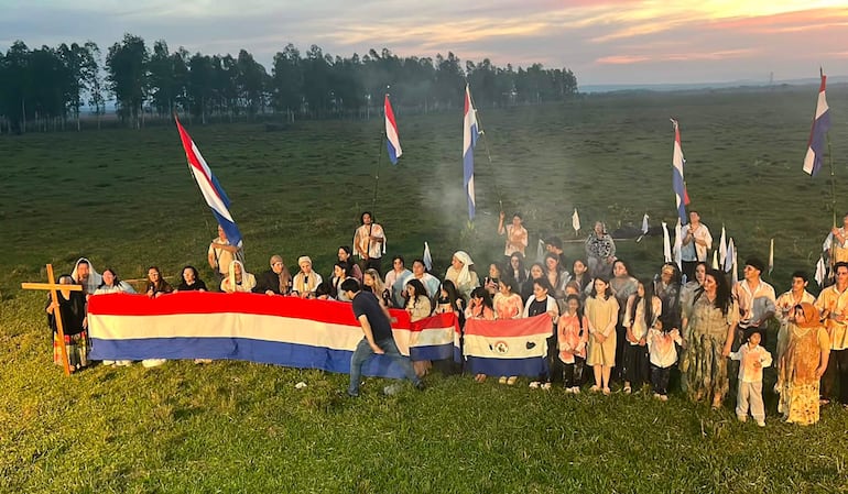 Numerosas personas sostienen una bandera de Paraguay en un acto, vestidos de ropa clara, con ambiente festivo y árboles al fondo.