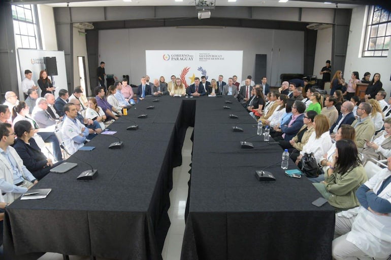 Asistentes en trajes formales y uniformes médicos escuchan a las autoridades en una mesa ovalada iluminada, con banner del Gobierno del Paraguay al fondo.