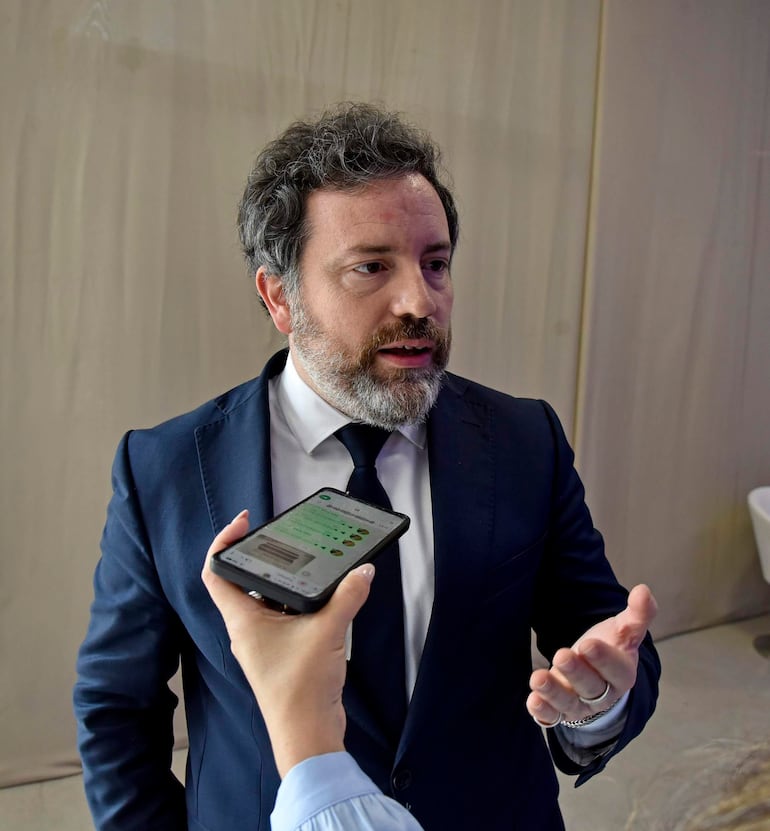 Hombre de barba y cabello castaño claro en traje oscuro, sosteniendo un teléfono mientras conversa en un ambiente neutro.