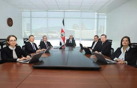 Seis adultos, dos mujeres y cuatro hombres, sentados en mesa formal con computadoras, en ambiente iluminado por luz natural, con bandera de Costa Rica de fondo.