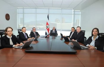Seis adultos, dos mujeres y cuatro hombres, sentados en mesa formal con computadoras, en ambiente iluminado por luz natural, con bandera de Costa Rica de fondo.