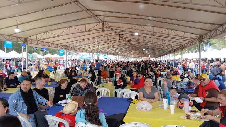 Gran cantidad de personas sonrientes en mesas bajo una carpa, algunos conversando y comiendo. Ambiente festivo con manteles azules y amarillos.