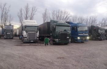 Estacionamiento con varios camiones de carga, silla verde en primer plano. Ambiente frío, árboles escasos al fondo.