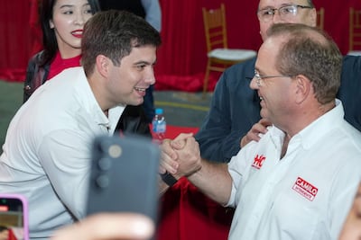 Camilo Pérez y Constantino Ayala sonríen y se saludan; fondo rojo con sillas de madera visibles.
