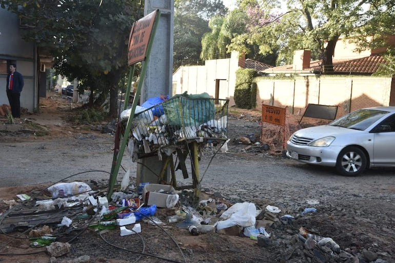 Persona con camiseta roja y abrigo oscuro observa un área deteriorada con basura y un auto plateado estacionado.
