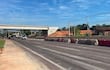 Carretera en construcción con vehículos circulando, barreras de cemento y un puente al fondo en un día soleado.