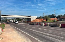 Carretera en construcción con vehículos circulando, barreras de cemento y un puente al fondo en un día soleado.