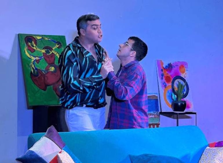 Hombre de camisa colorida y pantalones blancos gesticula animado, mientras otro con camisa a cuadros observa sorprendido en un escenario colorido.