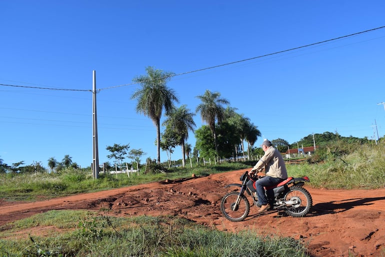 Hombre con camisa clara y sombrero, montando una motocicleta en un camino de tierra con vegetación de fondo.