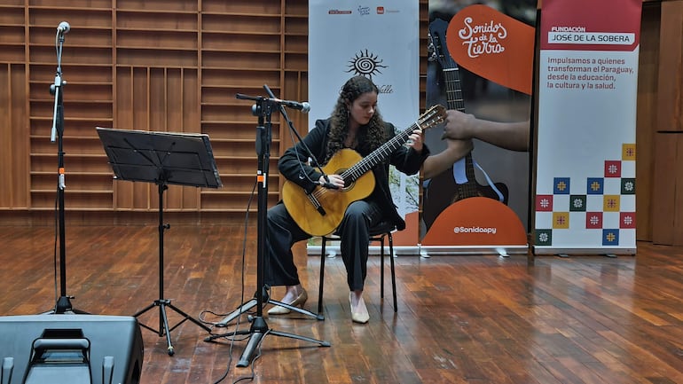 María Paz Cubells sentada, tocando guitarra clásica, con cabello rizado y conjunto negro, en un auditorio con micrófonos y pancartas de fondo.
