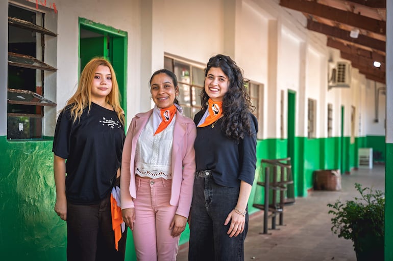 Sofía Hume, Thiara Pérez y Tamara Ruiz Díaz sonríen con pañoletas naranjas, en un pasillo con paredes verdes y blancas.