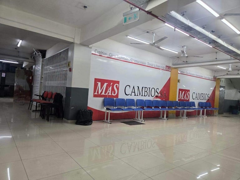 Interior de oficina de cambios con logotipo 'MÁS CAMBIOS S.A.', sillas azules y rojas, iluminado artificialmente y limpio.