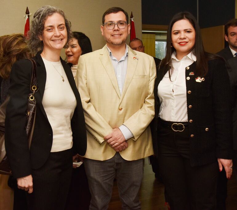 Mujer con blazer negro y blusa clara, hombre en chaqueta clara y camisa de cuadros, mujer en abrigo negro, todos sonrientes entre otros asistentes.