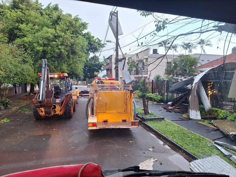 Retroexcavadora en acción y camión amarillo en calle con escombros y ramas tras tormenta en un vecindario residencial.