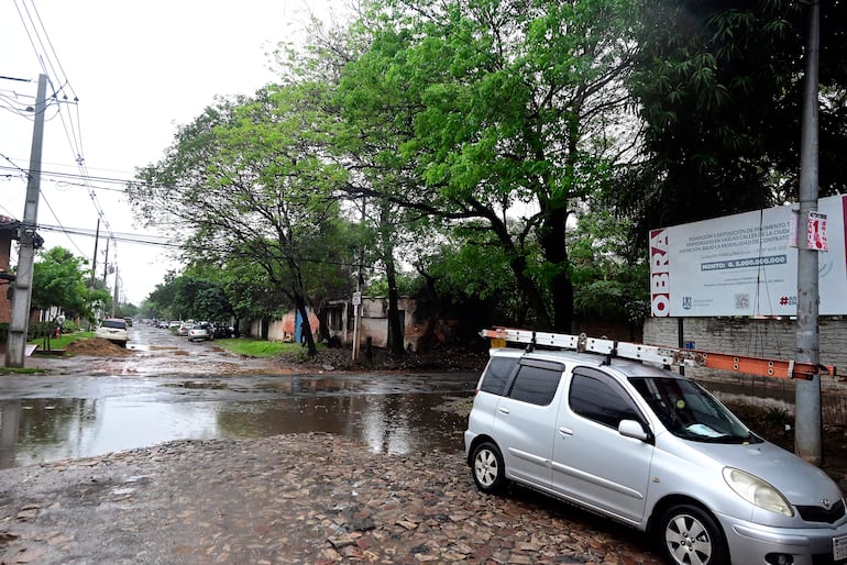 Calle empedrada y encharcada con un vehículo plateado estacionado y una obra en construcción al fondo.