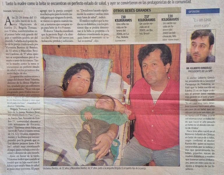 Mujer de cabello largo y oscuro sostiene a un bebé en brazos, mientras hombre con camisa roja observa con expresión tranquila.
