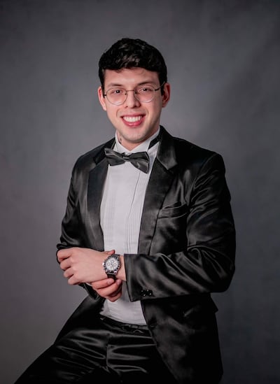 Joven con esmoquin negro y gafas sonríe a la cámara en un fondo neutro durante un retrato formal.