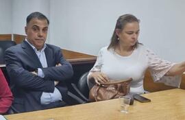 Javier Díaz Verón con traje oscuro y María Selva Morínigo en blusa blanca, conversan en mesa con un vaso y un móvil.