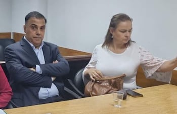 Javier Díaz Verón con traje oscuro y María Selva Morínigo en blusa blanca, conversan en mesa con un vaso y un móvil.