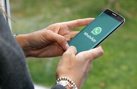 Manos de una mujer con un pulover gris y pulseras en las muñecas, que sostiene un teléfono celular abriendo la aplicación de WhatsApp.