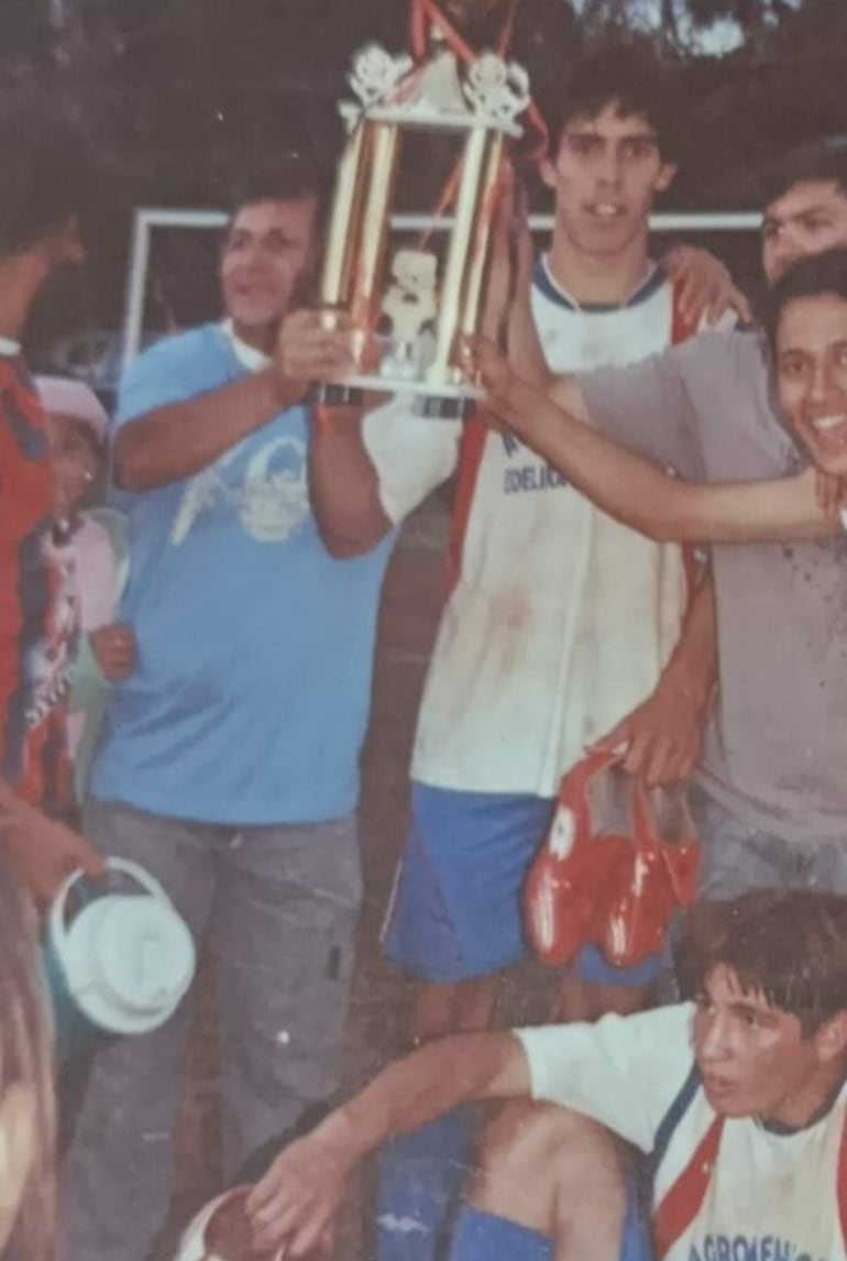 Gabriel Ávalos en camiseta blanca con detalles rojos y azules, levantando un trofeo con felicidad junto a cuatro personas en un campo de fútbol.
