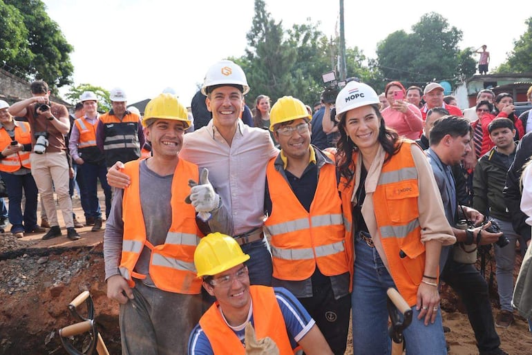 Santiago Peña y Claudia Centurión con cascos de seguridad sonriendo en la obra, rodeados de trabajadores y espectadores.