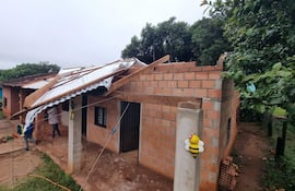 Paulina Contreras y un ayudante, vestidos casuales, reparan su casa de ladrillo con tejado dañado, rodeados de vegetación.