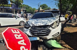 El automóvil blanco Hyundai está dañado y montado sobre un cartel de pare, con personas observando el accidente.