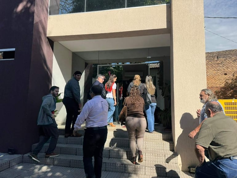 Grupo de personas, entre ellas una mujer de camiseta roja y un hombre con planta, ingresa a un edificio bajo luz solar.