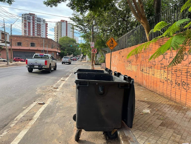 Vista de calle sin personas, con vehículos, contenedores de basura y graffiti en la pared.