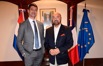 Hombre con traje gris y corbata de lunares junto a otro de blazer azul oscuro y barba, en un entorno con banderas de Paraguay y Francia.