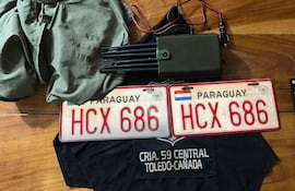 Un dispositivo rectangular verde oscuro junto a chapas de matrícula 'HCX 686' y prendas de la marca New Balance sobre mesa de madera.