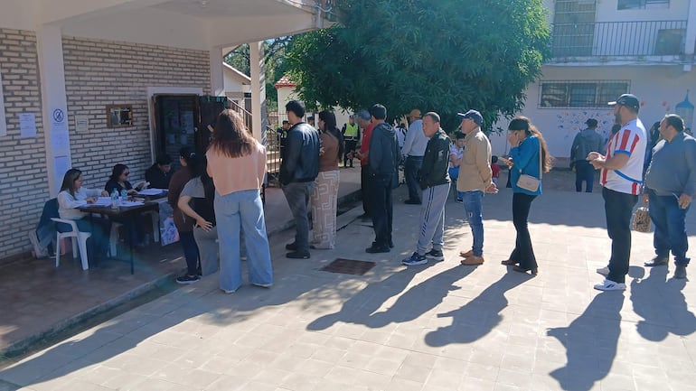 Grupo de personas en fila, algunas conversando, vestidas con ropa casual, en un ambiente de trámite administrativo al aire libre.