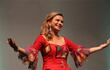 Rebecca Arramendi, sonriente y gesticulando, viste un vestido rojo con flores mientras actúa ante un público al fondo.