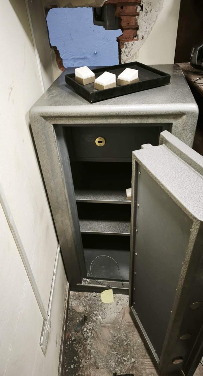 Caja fuerte abierta con objetos claros en bandeja negra, boquete en la pared y escombros en el suelo.