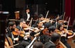 Violinistas en el centro, músicos en vestimenta formal tocan instrumentos en un auditorio iluminado.