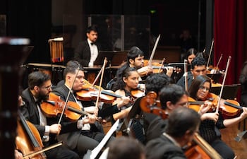 Violinistas en el centro, músicos en vestimenta formal tocan instrumentos en un auditorio iluminado.