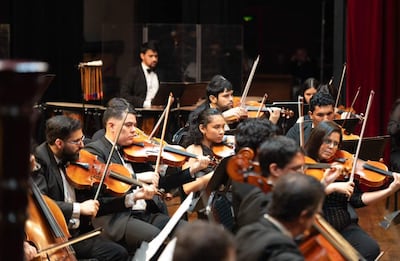 Violinistas en el centro, músicos en vestimenta formal tocan instrumentos en un auditorio iluminado.