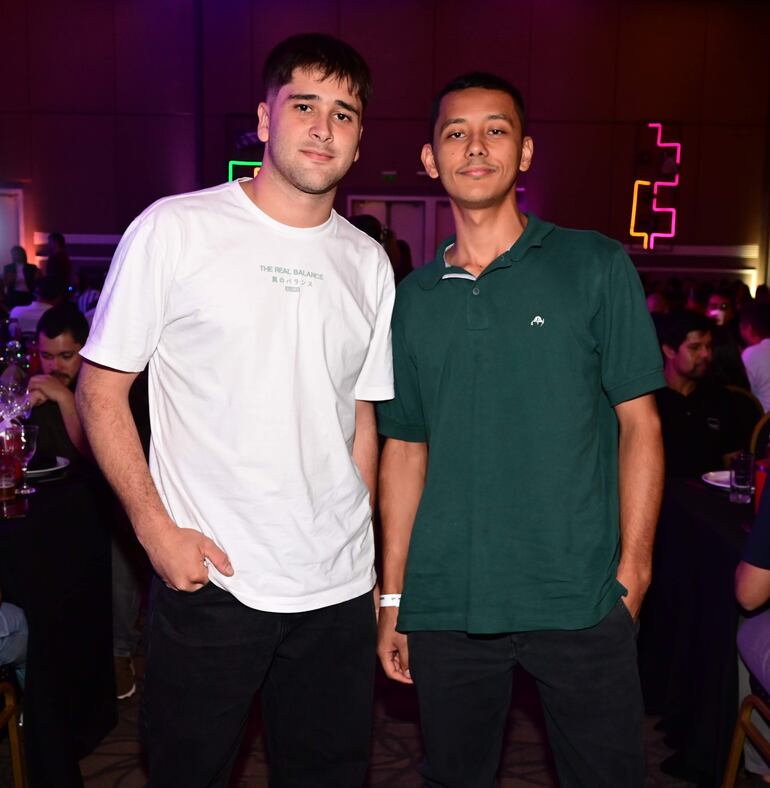 Dos hombres, uno con camiseta blanca y otro con camiseta verde, en un ambiente festivo con luces de colores y mesas al fondo.