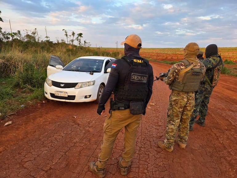 Agentes de la SENAD en chalecos tácticos y gorras, observan cuidadosamente un vehículo blanco en un entorno rural.