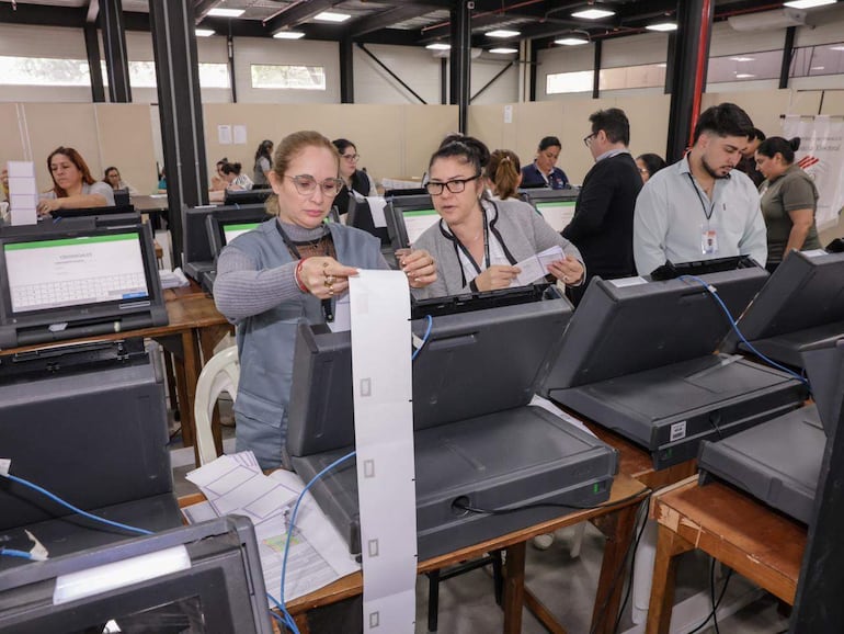 Mujer con gafas y cabello recogido revisa documentos, rodeada de otras personas en un ambiente de trabajo electoral.
