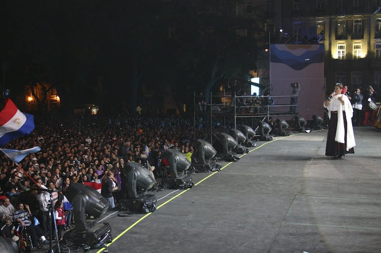 Lizza Bogado en abrigo blanco, hablando al público entusiasta, mientras se ven banderas y un ambiente festivo.