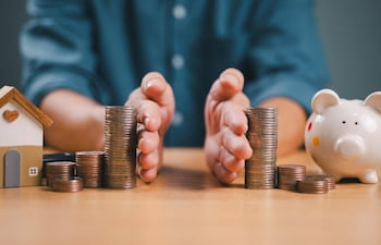 Mano humana sostiene monedas junto a alcancía en forma de cerdo y casa de juguete, simbolizando ahorro y finanzas.