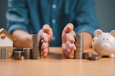 Mano humana sostiene monedas junto a alcancía en forma de cerdo y casa de juguete, simbolizando ahorro y finanzas.