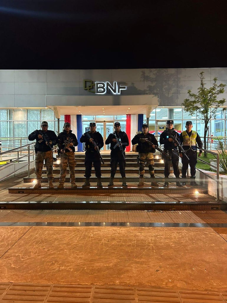 Siete policías armados en uniforme alineados frente a un edificio con cartel del BNF, en un ambiente nocturno.